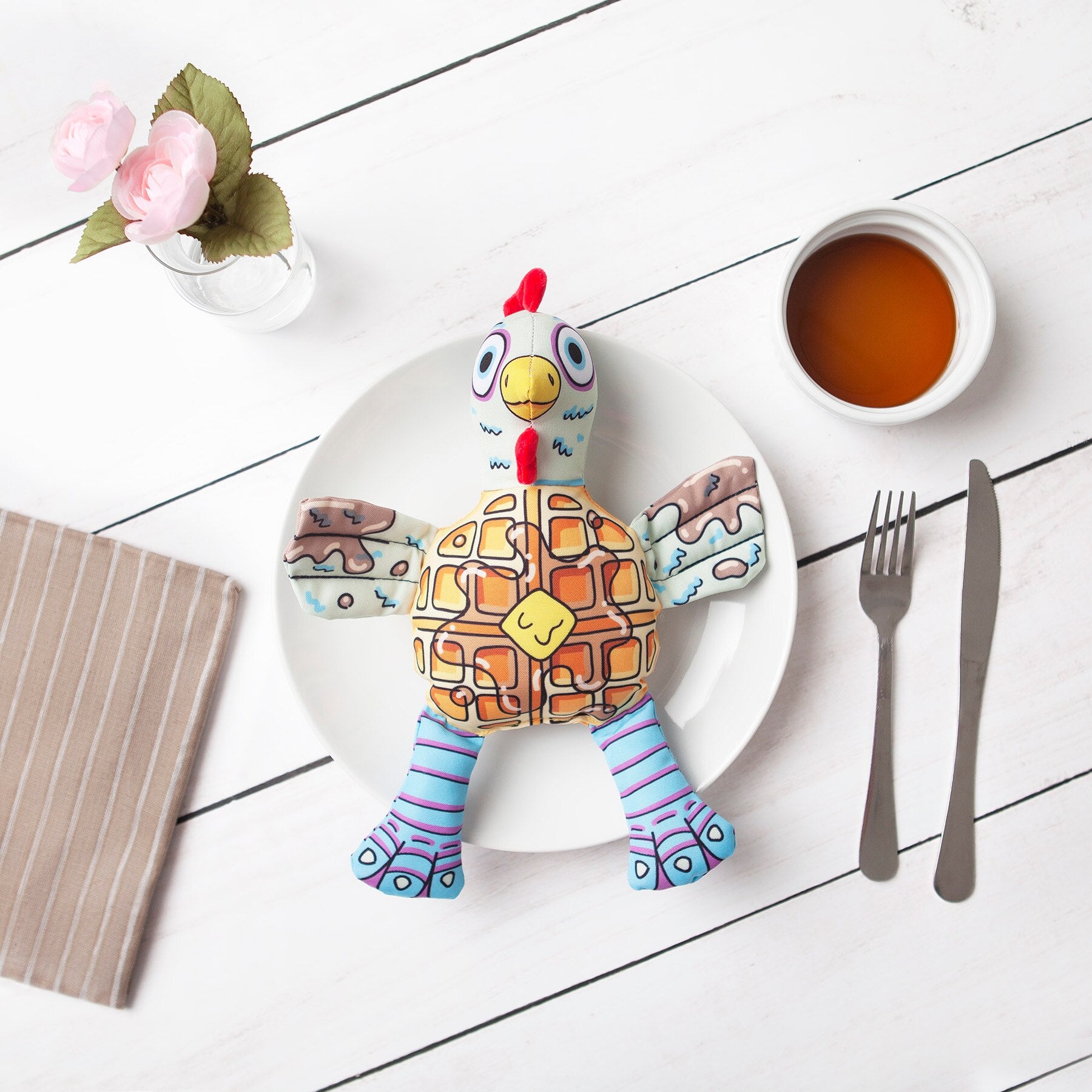 A FAT CAT Foodies Chicken n Waffles dog toy, with a waffle-patterned plush rooster, sits on a white plate on a wooden table. Nearby are cutlery, pink flowers in a glass, and a small jar of honey or syrup. This Fatcat toy perfectly displays floppability®.