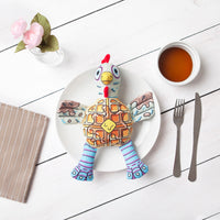 A FAT CAT Foodies Chicken n Waffles dog toy, with a waffle-patterned plush rooster, sits on a white plate on a wooden table. Nearby are cutlery, pink flowers in a glass, and a small jar of honey or syrup. This Fatcat toy perfectly displays floppability®.
