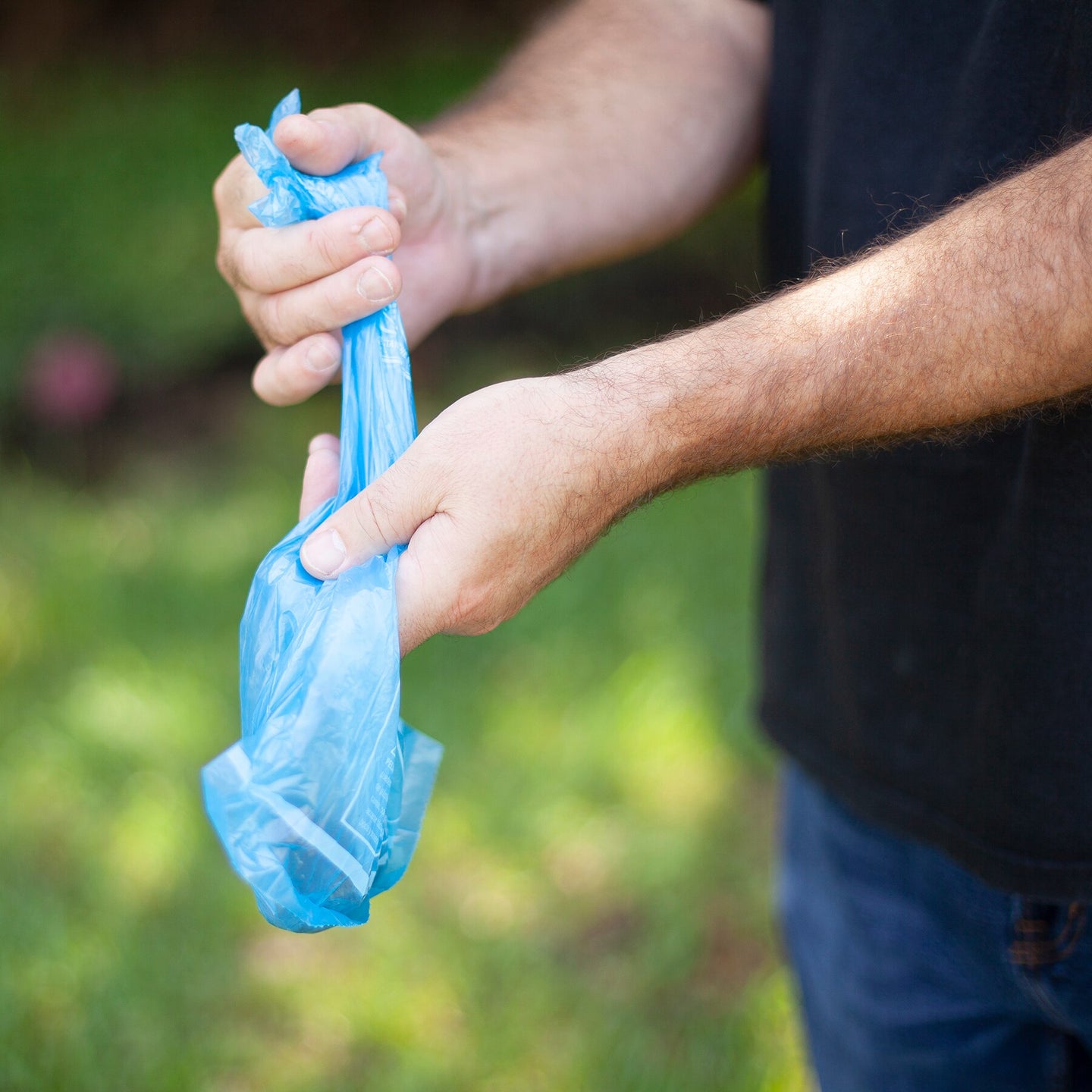 In a grassy setting, someone wearing a black shirt and blue pants uses their hands to tie an Arm & Hammer Easy-Tie Dog Waste Bag in blue, featuring activated baking soda for odor control.