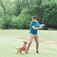 A man in a blue shirt and khaki pants playfully holds his Chuckit! Fetch Flight dog toy, facing a small brown dog. Theyre on a grassy field surrounded by trees, and the dog eagerly looks up at the durable frisbee by Chuckit.