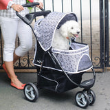 A fluffy white dog sits contentedly in a Gen7Pets Black Onyx Promenade Pet Stroller, adorned with a circular pattern and all-terrain wheels. The stroller is pushed by someone in white pants and orange shoes, with a black gate visible in the background.