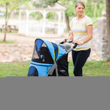 Wearing a yellow striped shirt and jeans, a woman pushes the Gen7Pets G7 Pet Stroller by Gen7 with its shock-absorbing wheels and blue cover through the park. She holds a cup in its holder, as trees and a gazebo blur softly in the background.