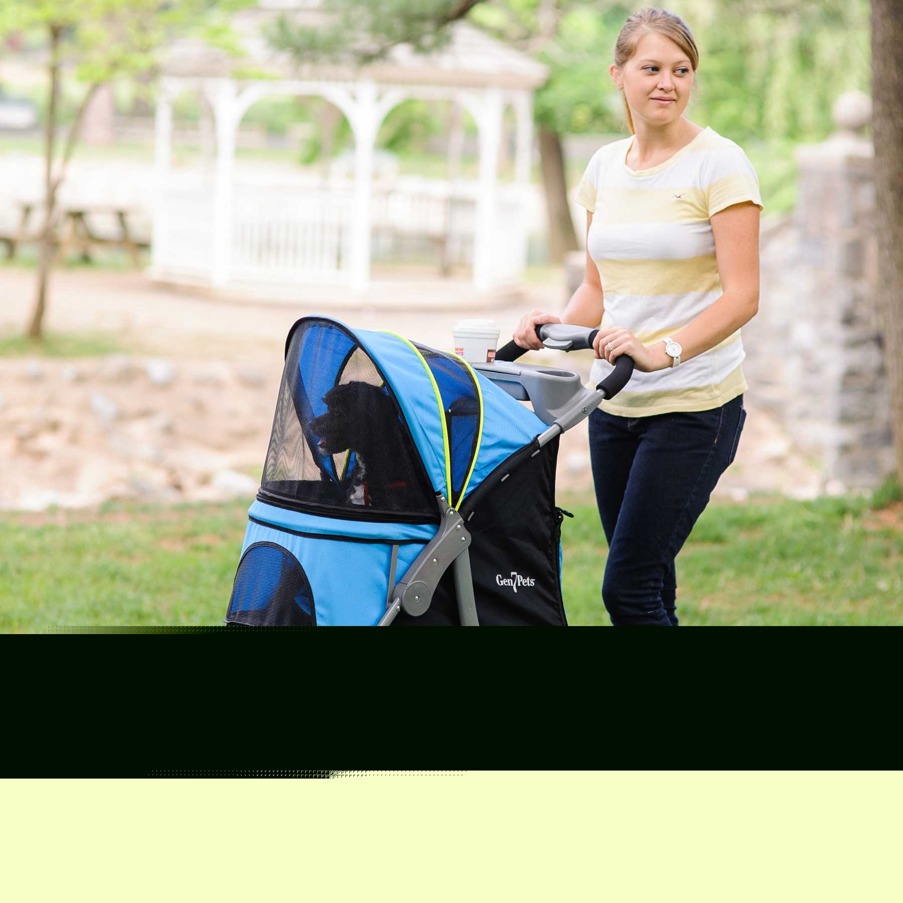 Wearing a yellow striped shirt and jeans, a woman pushes the Gen7Pets G7 Pet Stroller by Gen7 with its shock-absorbing wheels and blue cover through the park. She holds a cup in its holder, as trees and a gazebo blur softly in the background.