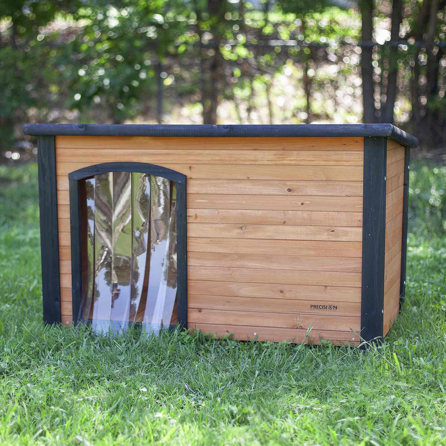 The Precision Pet Outback Dog House Door features a wooden structure with a flat black roof and weather-resistant clear plastic flap, set on a grassy lawn surrounded by trees, ensuring your pets comfort in any season.