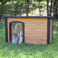 The Precision Pet Outback Dog House Door features a wooden structure with a flat black roof and weather-resistant clear plastic flap, set on a grassy lawn surrounded by trees, ensuring your pets comfort in any season.