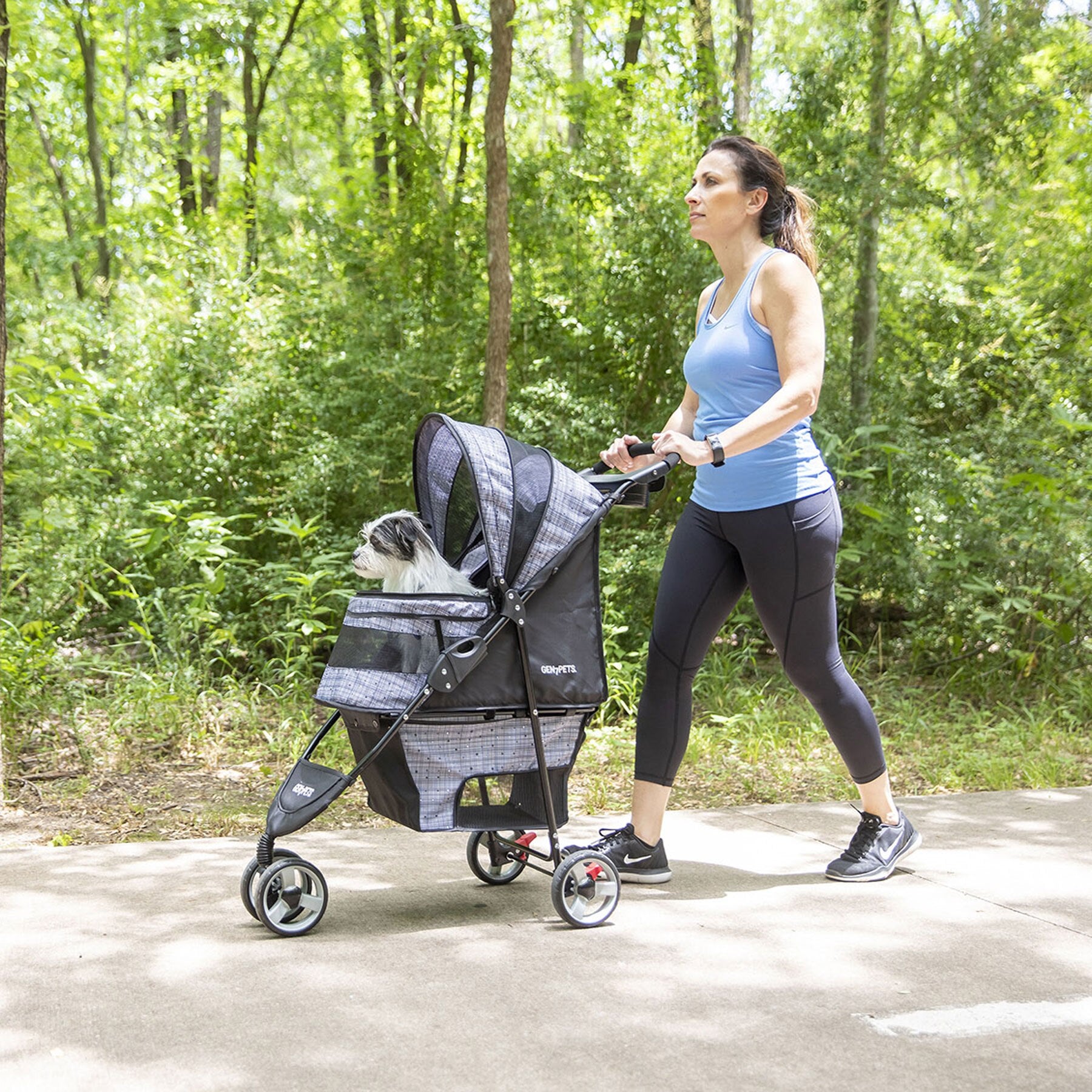 A woman in athletic wear energetically walks on a wooded path, pushing the Gen7Pets Starry Night Gray Regal Plus Pet Stroller. Inside, a small dog sits comfortably under the Smart-Canopy, enjoying the ride as its owner focuses on her journey.