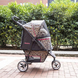 A small dog relaxes in a Gen7Pets Cheetah Promenade Pet Stroller, featuring a Smart-Comfort Pad and safety tethers, under a transparent cover. Its parked on a brick path, surrounded by green bushes and with a brick building visible behind.