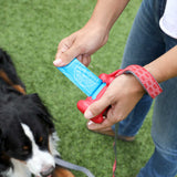 A person holds a blue waste bag from the red Arm & Hammer Bone Dispenser as a black and white dog sits on the grass. The person, in jeans and a patterned wristband, appreciates the odor control of the Arm And Hammer dispenser.