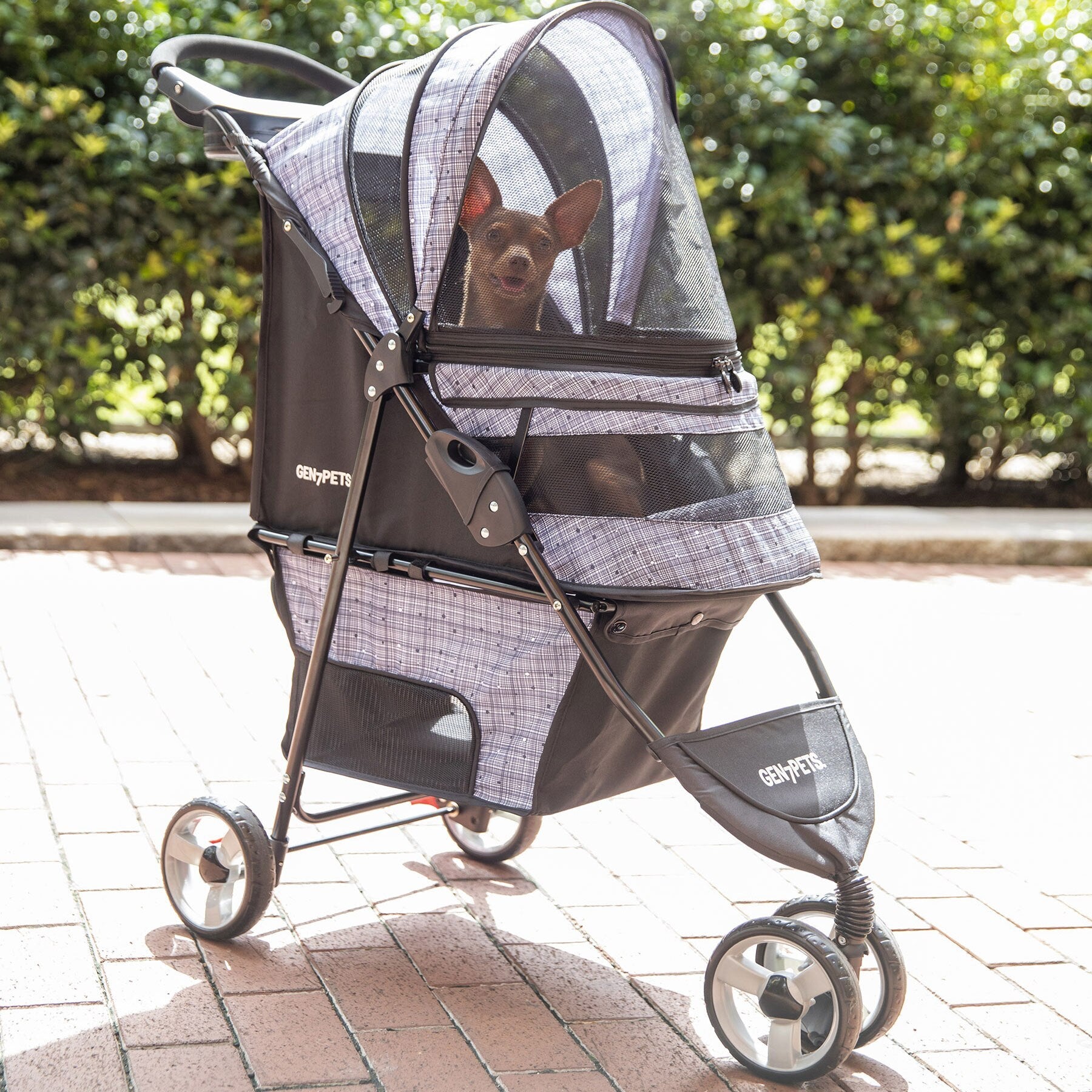 A dog relaxes inside a Gen7Pets Starry Night Gray Regal Plus Pet Stroller featuring a Smart-Canopy and mesh cover, parked on a brick path, with sunlight-blurred bushes in the background.