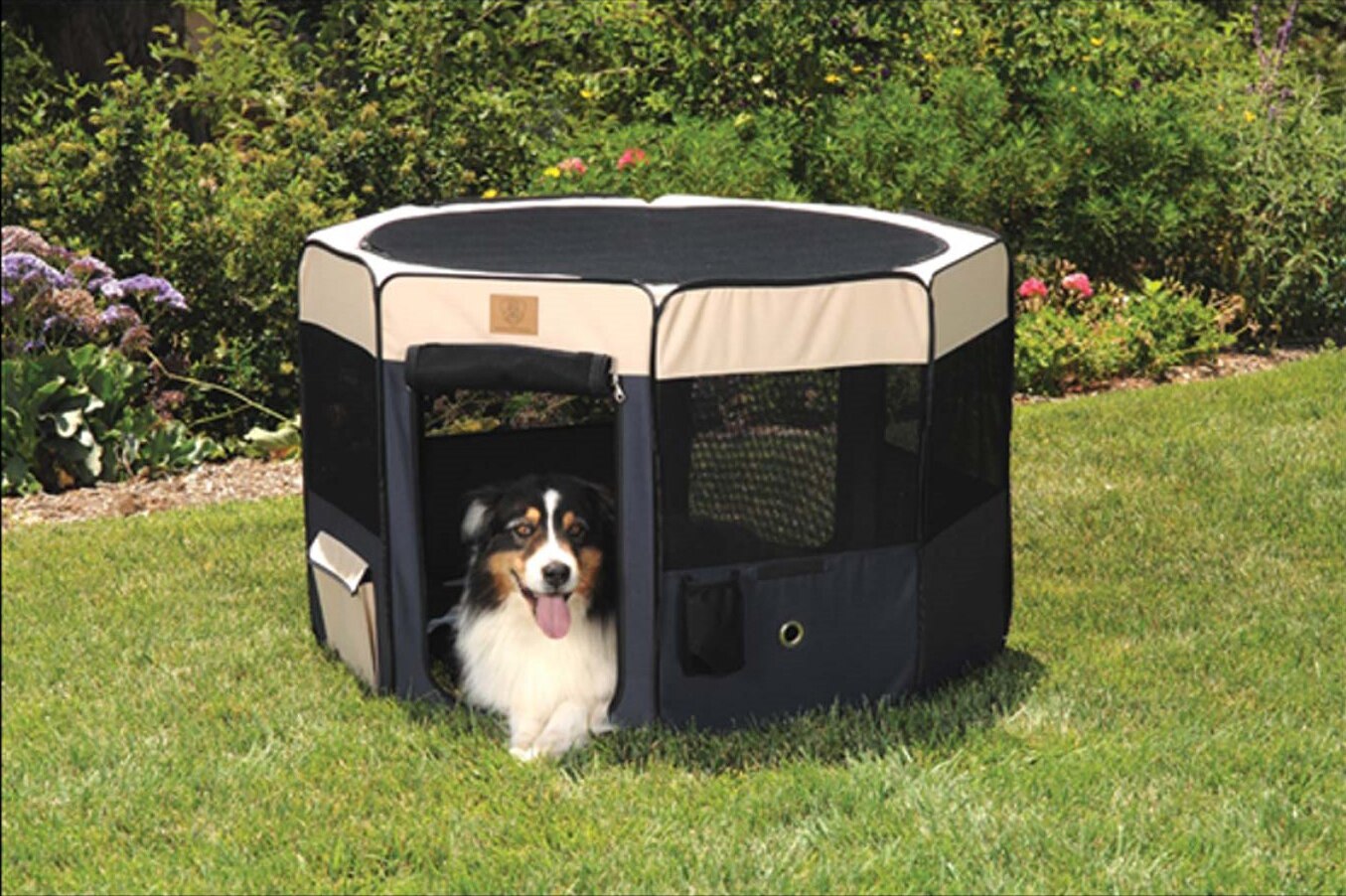 A dog with a black and brown coat and a white face sits inside an open Precision Pet Soft Side Play Yard on a grassy lawn, set against lush garden bushes and vibrant flowers. This water-resistant accessory is from Precision Pet Products.