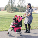 A woman in a gray dress and black boots strolls along a path with her Gen7Pets Raspberry Sorbet Regal Plus Pet Stroller, featuring the Smart-Canopy. A small black dog enjoys the ride, surrounded by lush green grass and trees.
