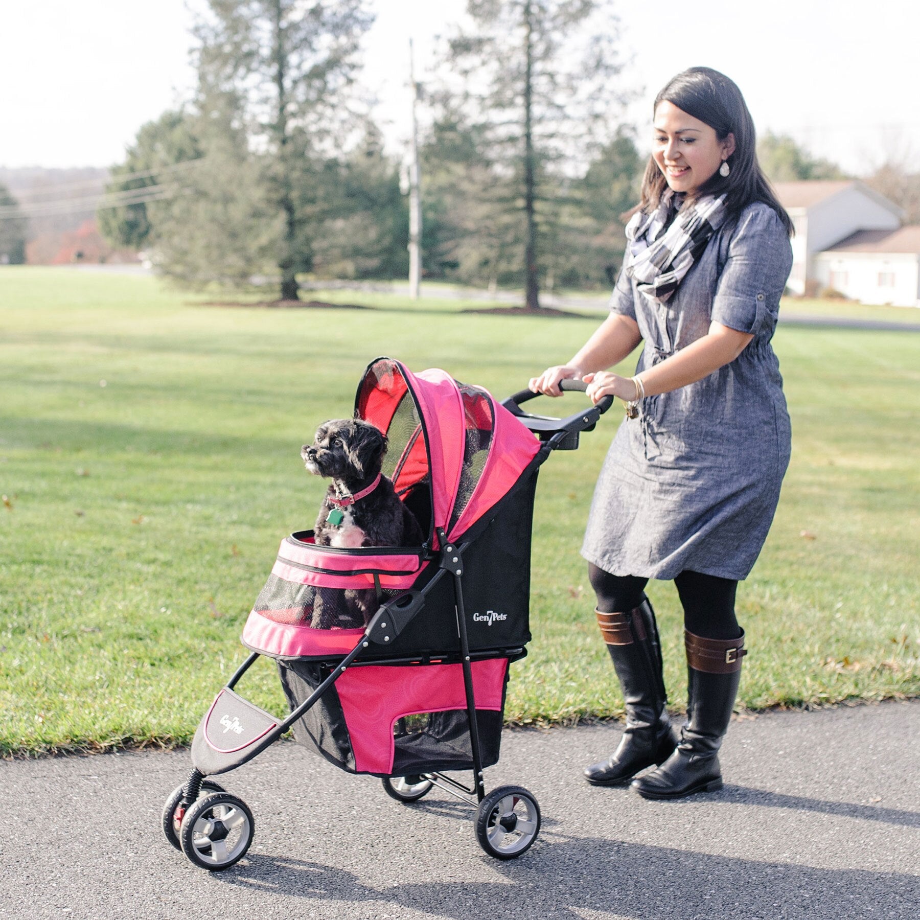 A woman in a gray dress and black boots strolls along a path with her Gen7Pets Raspberry Sorbet Regal Plus Pet Stroller, featuring the Smart-Canopy. A small black dog enjoys the ride, surrounded by lush green grass and trees.