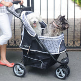 Two small dogs, one white and fluffy, the other gray and shaggy, sit comfortably in a Gen7Pets Black Onyx Promenade Pet Stroller with Smart-Comfort Pad. A person in white pants pushes it effortlessly using its All-Terrain Wheels as a black gate stands gently behind.