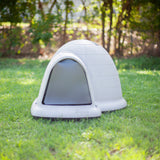 A white plastic dog house from the Petmate Indigo Door collection sits on a green lawn. Trees and a fence in the blurred background provide shade, creating an ideal setting for the Petmate Outdoor Shelter experience.
