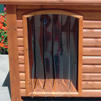 This weather-resistant wooden doghouse, set against concrete and vibrant orange flowers, boasts a Precision Pet Products Outback Dog House Door. Its natural wood finish and peaked roof offer an inviting, cozy shelter for your furry friend.