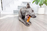 A gray dog lies on a wooden floor, happily chewing a Wild Eats Salmon Flavored Collagen Retriever Adult Roll. In the background, white stairs with storage drawers and a large potted plant are visible in the bright natural light.